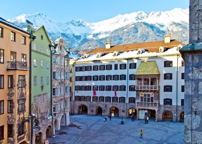 Farbenfrohe europäische Gebäude und ein Platz in der Nähe des Appartments Sellrain mit schneebedeckten Gipfeln im Hintergrund.